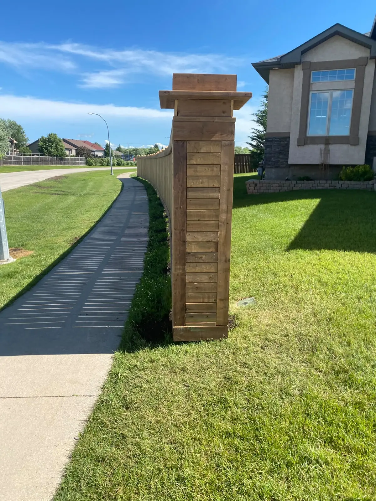 Long wood privacy fence along a sidewalk, ending in a clad wood pillar with a cap